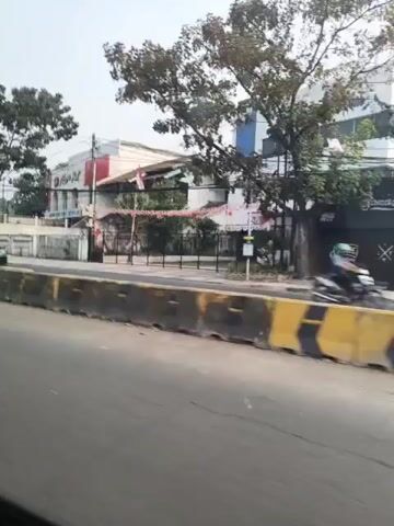 The video captures a street scene, likely in an urban or suburban area. The perspective is from a moving vehicle, suggesting a journey or commute. The road is visible in the foreground, with a concrete barrier separating it from the sidewalk or median. Beyond the barrier, there's a sidewalk area with some greenery and trees lining the street. Several buildings are visible in the background, appearing to be a mix of commercial establishments and possibly residential properties. One building has a red and white facade, and another has awnings, suggesting businesses. A few vehicles, including a black SUV and a white car, are present on the road or parked nearby. There are also a couple of motorcycles visible. The sky is overcast, indicating a cloudy day. The overall impression is of a normal day on a street with moderate traffic and commercial activity.