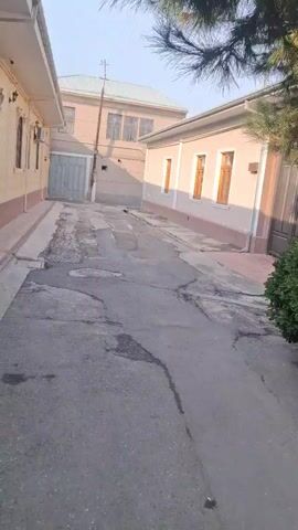 The camera films a narrow street with cracked asphalt between low buildings, under a light blue sky. The buildings are mostly one-story, though one is two stories, and painted in light, neutral tones. All the buildings have windows with wooden frames. The street shows signs of water damage and wear.  A utility pole stands near the two-story building, and a metal manhole cover is visible in the middle of the street. There are no people visible in the scene.