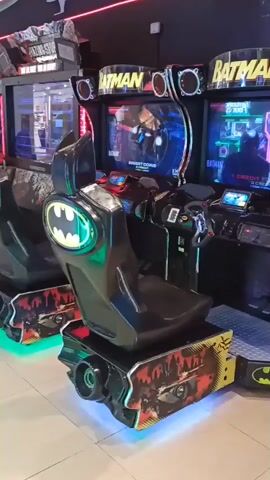 This video shows two Batman: The Brave and the Bold arcade machines, likely located in an arcade or entertainment center. The machines are designed to resemble the Batmobile, with a steering wheel, a seat, and a screen displaying game graphics. The screens show gameplay footage, with the title "BATMAN: THE BRAVE & THE BOLD" clearly visible. The arcade machines are lit with neon lights, and the overall atmosphere appears to be that of a vibrant entertainment venue. The video appears to be shot indoors, with artificial lighting. There is no indication of the time of day or weather conditions. The location is identified as Modugno, Italy.