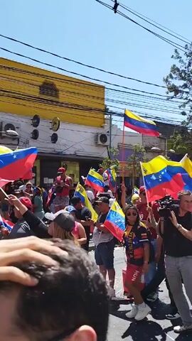 The video shows a lively street demonstration. A group of people is marching down the street, waving Venezuelan flags of varying sizes. Some participants wear the colors of the Venezuelan flag in their clothing. A professional cameraman films the procession. People wear sunglasses, hats, and other casual attire. The background features a bright yellow building with signage, power lines overhead, and trees along the street. The atmosphere appears to be celebratory and energetic.