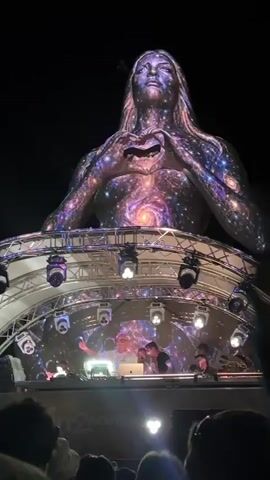 A nighttime outdoor music festival scene. A large inflatable or projection of a human figure, with long hair and hands forming a heart shape over its chest, is displayed prominently in the background. The figure is illuminated with swirling, cosmic-like light patterns. Below this, a stage structure with multiple spotlights and speakers is visible. On the stage, several individuals, presumed to be DJs, are operating equipment. In the foreground, a crowd of people is gathered, looking towards the stage. The overall atmosphere suggests a lively event with a focus on visual spectacle and music.