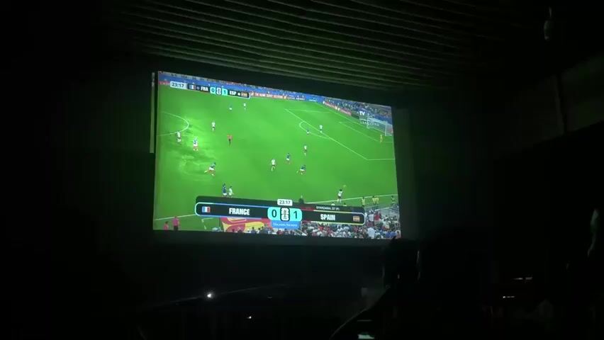 A projected image of a soccer match is displayed on a large screen in a dimly lit room. The screen shows a live game with players in white and blue uniforms on a green field. A scoreboard at the bottom indicates FRANCE 0 and SPAIN 1. The time displayed is 23:19. In the foreground, silhouettes of people are visible watching the screen. The room has a textured ceiling. The atmosphere appears to be that of a sports bar or a viewing party.