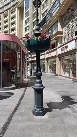 The video shows a street scene with several buildings and a miniature replica of the Leaning Tower of Pisa. In the foreground, a black lamppost with a flower basket is visible. To the left, a pink building with the word "PANDORA" and its logo is present, featuring a circular design with a crown on top. Next to the Pandora store, the miniature Leaning Tower of Pisa stands enclosed by a black fence. In the background, other buildings with intricate architectural details can be seen, along with shopfronts. The street is paved with gray tiles, and there is a drainage grate on the left side of the frame. The lighting suggests it is daytime, with shadows cast by the objects. No people are clearly visible in the frame.