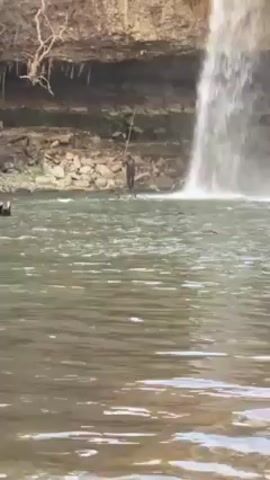 A distant figure stands in the water at the base of a waterfall. The waterfall is tall, with water cascading down from a rocky overhang.  The water body in front of the waterfall is still, with some ripples on the surface.  Icicles hang from the rock overhang.  The environment appears to be a natural area with trees visible at the edge of the frame.