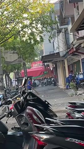 A person in a blue shirt is sweeping the sidewalk in front of several parked motorcycles. In the background, there are more motorcycles and buildings. The sun is shining and trees are visible.