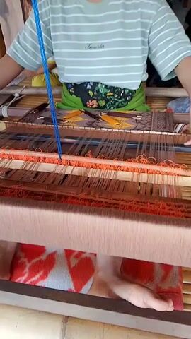 A person is shown sitting and weaving on a loom. The person wears a light green t-shirt with white horizontal stripes and a black and green patterned apron. Their feet are visible, wearing a red and white patterned cloth. The loom is made of wood and bamboo. The weaving is done with orange and brown threads, creating a patterned fabric. The person is actively engaged in the weaving process, using their hands to manipulate the threads and shuttle. The environment appears to be indoors, with other people and objects partially visible in the background.