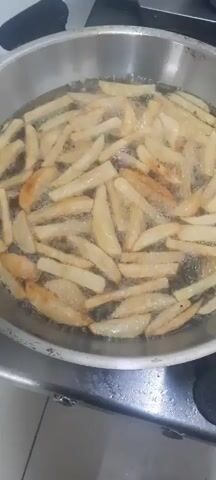 A close-up, overhead shot shows french fries being deep-fried in a large metal pot filled with bubbling oil. The fries are mostly golden brown, with some lighter and darker pieces scattered throughout. The oil is actively bubbling, indicating a high temperature. The pot is placed on a stovetop, suggested by the metallic surface beneath it and a hint of a black control knob to the right. A metal mesh skimmer is briefly lowered into the oil from the upper right corner of the frame, suggesting the fries may be close to being done or are being stirred. The lighting is somewhat dim, creating a slightly grainy image quality, and the overall setting appears to be a kitchen.