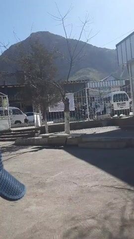 The video shows a young girl walking towards the camera. She has brown hair pulled back in a ponytail and is wearing a white long-sleeved shirt with black stars and black pants. The girl is on a paved area, and behind her is a building with a fenced-in area. In the background, there is a mountain, a car, and some trees.