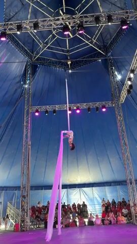 A female aerialist performs a routine on a silk fabric suspended from a ceiling structure within a blue tent. She is wearing a light-colored costume and her hair is dark brown. The silk fabric is white at the top and transitions to a vibrant pink as it hangs down. The aerialist is positioned high above a seated audience, some of whom are visible in the background. The tent is illuminated by pink and green stage lights. Metal scaffolding supports the tent structure and the rigging for the aerial performance.