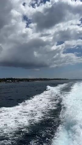The video shows a view from a boat moving through the ocean. The water is dark blue and choppy, with a white wake trailing behind the boat. In the distance, a shoreline is visible with palm trees, houses, and several small boats. The sky is partly cloudy with patches of blue sky and dark clouds. The sun appears to be shining, as indicated by the bright reflections on the water and the light illuminating the clouds. The overall impression is a tropical coastal scene.