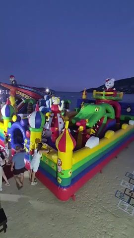 A drone shot begins by looking down at a large, inflatable play structure on a sandy beach. The play structure is colorful, with red, yellow, green, blue, and purple sections. It features a large green crocodile with its mouth open, a slide, and several towers resembling onion domes. A white teddy bear wearing a Santa hat sits on top of the crocodile. Several people are visible near the play structure. Two people, a man and a woman, are on the left side of the frame, and a woman in a white dress is interacting with the play structure. In the background, the ocean and a hilly coastline are visible under a dark blue twilight sky. A hopscotch grid is drawn on the sand to the right of the play structure.