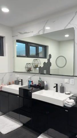 The video shows a well-lit modern bathroom with a double sink vanity, dark cabinets, and a large rectangular mirror. The mirror reflects the back of the bathroom, where a toilet, hamper, and small round mirror are visible.  A woman appears in the mirror, holding a phone and taking a selfie. She raises her arms and appears excited. She is wearing a black sweatshirt and pants.
