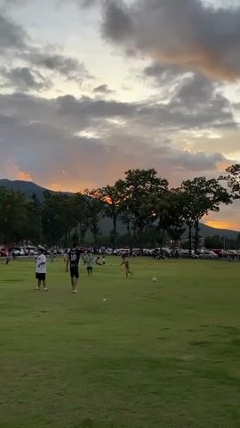 The video shows a wide shot of a grassy field at dusk. Several people are scattered across the field, some of whom appear to be playing soccer. The sky is cloudy with streaks of orange and yellow indicating a sunset. Trees line the background, and behind them are mountains. Cars are parked in a lot behind the trees. The lighting suggests it is late afternoon or early evening.