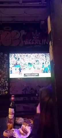 The video begins with a close-up of a television screen showing three men in white jerseys with blue stripes, smiling and hugging. One man on the left appears to be speaking. The television is mounted on a wall decorated with graffiti that says "BEERLIN" in large letters. Below the TV, a bottle of red wine and other items are visible on a surface. As the video progresses, the camera pans upwards and to the right, revealing a dimly lit interior with purple and blue lighting. More graffiti is visible on the wall. The shot then expands to show a group of people in the foreground, their backs to the camera, looking towards the television. In the background, the scene opens up to a wider view of what appears to be a bar or entertainment venue with more people and colorful lighting, including a prominent vertical blue light. The overall environment appears to be lively and social, with patrons watching something on a screen.