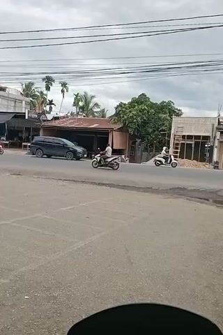 The video shows a street scene with several motorcycles and a dump truck. The motorcycles are moving in various directions, and some are carrying passengers. A black minivan is parked on the side of the road. Buildings line the street, some appearing to be shops or businesses. Overhead power lines are visible against a cloudy sky. The road surface is asphalt, with painted lane markings. The overall impression is a typical street scene in a populated area, likely during daylight hours.