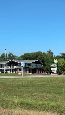 The video shows a motel with a large sign that says "All Star" and shows a green star. The motel has two stories and an outdoor walkway. There are cars parked in front of the motel and a flagpole with a Canadian flag. In the background, there are trees and a clear blue sky. There is also a road in the foreground of the video with a traffic light. The camera pans across the scene, showing different angles of the motel and its surroundings.