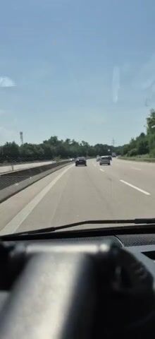 The video captures a view from the interior of a car driving on a highway. The perspective is from behind the dashboard, with the windshield in focus. Through the windshield, we see a multi-lane highway with several cars ahead. The sky is bright blue, indicating a clear day, likely daytime given the lighting. Lush green trees line the sides of the highway, suggesting a rural or suburban setting. The road appears to be in good condition.

The primary activity is driving on a highway, likely in Germany, given the "Malsch, Germany" location context. The cars visible are modern passenger vehicles. The mood is neutral, simply documenting the experience of highway travel. There are no discernible emotions or specific events beyond the act of driving. The image is clear and shows good visibility, without any obstructions. There is no visible text in the frame.