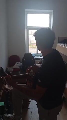 A young man wearing a black t-shirt and grey pants plays an acoustic guitar in a dimly lit room. He is seated and facing away from the camera, looking towards a window. The room contains a bunk bed with bedding, a red chair, and a black gaming chair. Clothes are draped over the chairs and on the floor. Sunlight streams through the window, illuminating the room.