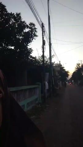 The video shows a dimly lit street scene at dusk or dawn. A single street lamp illuminates the left side of the road. Trees and houses line the street, with power lines crisscrossing overhead. The sky is a soft gray with hints of orange and yellow from the setting or rising sun. The road is asphalt and appears to be a residential area. There are no visible people or distinct actions taking place. The camera appears to be moving slightly, suggesting it is being held or is on a moving vehicle.