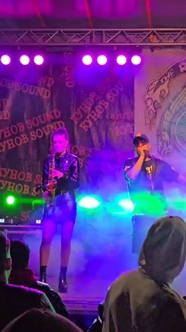 A female saxophonist and male vocalist are performing on a stage lit with purple and green lights. The stage backdrop is covered with repeated text, “Kyhob Sound,” and a logo for “The Riders MC.” The saxophonist, in a black outfit, stands to the left, while the vocalist, also in black, gestures with a microphone in his hand on the right. A hazy fog fills the stage. Audience members are seen in the foreground, some with their arms raised.