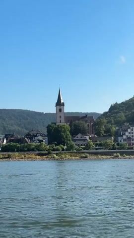 This is a video that pans across a scenic riverfront view of the town of Lorch am Rhein, Germany. The foreground is dominated by the rippling, blue-green water of the Rhine River. Beyond the river, a picturesque town nestled at the base of a lush, tree-covered hillside is visible.

Dominating the town's skyline is a prominent church with a tall, steeple. Several other buildings, with characteristic European architecture, are clustered together along the riverbank. The hillside behind the town is a vibrant green, dotted with trees and some visible terraced vineyards, indicating a rural or semi-rural setting. Near the top of the hill, the ruins of a stone tower or small castle can be seen.

The scene suggests a clear, sunny day with a bright blue sky. The water appears calm, indicating gentle movement, possibly from a boat or a slight current. The overall atmosphere is peaceful and idyllic, capturing the beauty of a German riverside town. No people are clearly visible, and there is no visible text in the video. The time of day appears to be late morning or early afternoon, given the bright sunlight and the shadows.