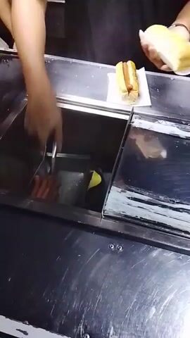 A person is making a hot dog at a hot dog cart.  The person has their right hand out of frame holding a hot dog in a bun on a paper wrapper. The person uses tongs to remove a hot dog from a container and place it in another bun.  They then close the lid to the container and use a spoon to wipe the lid clean.  The hot dog cart is made of stainless steel.