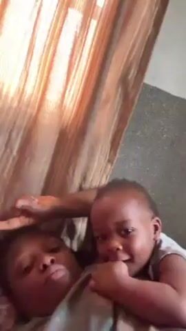 A woman and a baby are lying down indoors. The woman is on her back with her eyes closed, and the baby is lying on top of her chest. The baby is looking at the camera and has a hand in its mouth. There is a brown curtain to the left of the frame and a gray wall to the right. The video appears to be taken at an upward angle.