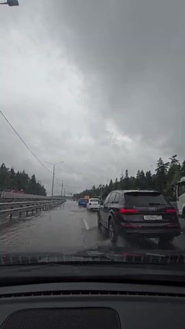 A rear view of a car driving on a wet highway during overcast weather. Cars are in front of the vehicle, including a black SUV and a white truck. The road is wet and appears to have puddles. Trees line the right side of the highway. The sky is heavily clouded and grey.