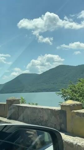 A brief video clip from a moving vehicle captures a scenic landscape. The view is from a car window, with a reflection of a hand holding a phone visible in the bottom left. Initially, a stone wall and a small structure are in the foreground. As the vehicle proceeds, a yellow metal bridge railing with black vertical stripes appears in the foreground, obscuring the stone wall. Beyond the railing, a large body of water stretches towards a backdrop of rolling, tree-covered green mountains. The sky is bright blue with scattered white cumulus clouds. Sunlight creates a noticeable lens flare in the upper right portion of the frame.
