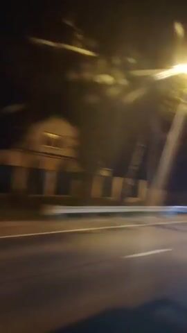 The video shows a blurry nighttime view from a vehicle moving along a road. Streetlights and trees are visible on the side of the road, with some lights creating bright streaks due to motion blur. A horizontal red light streak indicates another vehicle passing in the opposite direction. The road surface is asphalt, and there is a white line marking the lane. The overall impression is of traveling at night, with the environment illuminated by artificial light sources.