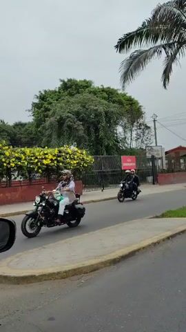 The video shows a group of motorcyclists riding down a street. They are all wearing helmets and riding in the same direction.  The street is lined with trees and buildings.  A sign for "Emergencia" (Emergency) is visible near a fenced area.  There is also a tall palm tree on the right side of the screen.  The weather appears cloudy and overcast.