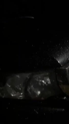 The video shows a close-up, low-angle shot of a black car at night, possibly in the rain. Water droplets are visible on the car's hood and fender. The headlights are on, illuminating the front of the car. The grille of the car is visible on the right side of the frame. The lighting is dim, and the background is dark, suggesting an outdoor setting at night.