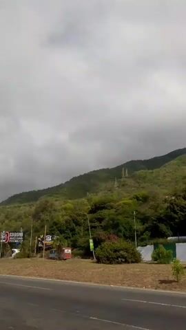 A vertical video shows a road with a slight curve to the right. On the right side of the road, there is a lush green hillside covered in trees and vegetation. Scattered on the hillside are various signs, including a large billboard with red and white lettering that appears to be an advertisement. Power lines and poles are visible on the upper part of the hill. On the left side of the road, there is a tall, multi-story building with a white facade and numerous windows. Cars are driving on the road, and there are some smaller trees and bushes along the roadside. The sky is partly cloudy with a mix of white and gray clouds. The video appears to be recorded from a moving vehicle.