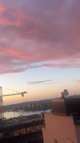 The video begins with a wide shot of a sky filled with pink and orange clouds, likely during sunset. In the distance, a construction crane stands tall against the horizon. As the video progresses, the camera pans to focus on a building with balconies, then shifts to capture people sitting at tables outside a restaurant or cafe. The atmosphere appears relaxed, with patrons enjoying the evening. The scene is illuminated by the soft glow of the sunset and some artificial lighting from the establishment. The camera then zooms in slightly on a television screen displaying what appears to be a sports event or broadcast, positioned above the outdoor seating area. Several people are visible, some facing the screen, others engaged in conversation. The overall impression is of a pleasant urban evening setting, with people socializing outdoors under a colorful sky.