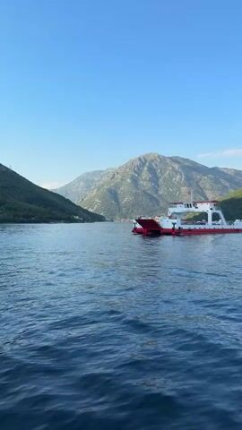A red and white ferry boat travels across a body of water. The ferry has a raised upper deck. The water is calm and reflects the clear blue sky. In the background, green, tree-covered mountains rise from the water's edge, and the peaks are covered in scrub and rock. The sun is high in the sky, casting a bright light on the scene.
