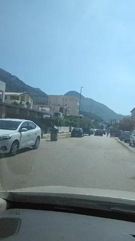 The video shows a view from inside a vehicle looking out at a street. The street is lined with parked cars and buildings. There are a few people walking on the sidewalk. The sky is blue and clear, and the sun is shining. There are mountains in the background. The video is of low quality and appears to be a screen recording.