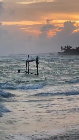 This media file captures a serene ocean scene during a dramatic sunset in Kathaluwa, Sri Lanka. The sky is a canvas of warm oranges and yellows, with dramatic clouds adding depth and texture as the sun dips below the horizon. A rough sea churns with white-capped waves rolling towards a sandy beach, suggesting a windy or choppy condition.

In the foreground, partially submerged in the turbulent water, stands a rustic wooden structure, possibly an old fishing platform or a unique piece of coastal art. Further out, a distant headland is silhouetted against the vibrant sky, dotted with palm trees and a small building with a lookout point, hinting at a tropical, coastal dwelling or resort. The overall atmosphere is one of tranquil beauty, with the powerful force of nature represented by the rough sea and the soft glow of the sunset creating a captivating contrast. The time of day is clearly sunset, with the lighting and cloud formations characteristic of this period. The location is identified as Kathaluwa, Sri Lanka, a detail that aligns with the tropical vegetation and coastal environment depicted. There is no visible text in the media file.
