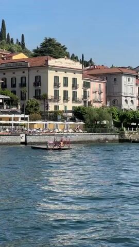 The video captures a bright, sunny day on a body of water, likely a lake, with buildings lining the shore in the background. The buildings are multi-story, painted in various pastel colors, and appear to be hotels or residences. One building prominently displays the text "HOTEL FLORENCE". A waterfront promenade with tables and umbrellas is visible in front of the buildings. On the water, a small, flat-bottomed raft with several people on it is moving. In the foreground, a dark-hulled motorboat with an orange interior and a canopy is cruising past, creating a wake. There are at least three people visible on the motorboat, one steering. The water is blue and choppy, reflecting the sunlight. The overall scene suggests a lively, tourist-oriented location.