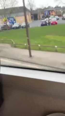 The video shows a view from inside a moving bus.  The camera is focused on the roadside seen through the window. A Lidl supermarket is visible across the road, with its parking lot holding several cars.  A few pedestrians are walking on the sidewalk. The weather appears overcast.  The bus passes by a bus stop sign painted on the road.