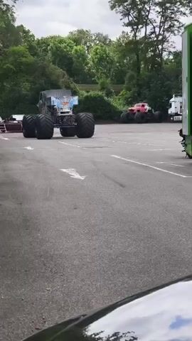 A monster truck with a blue and white design is on a paved surface. In the background, a red monster truck is visible. A person runs across the paved surface in front of the blue and white monster truck. The environment appears to be an outdoor area with trees and foliage in the background. A white truck and a green trailer are parked on the right side of the frame. The perspective is from inside a vehicle, with a black hood visible in the foreground.