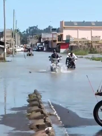 The scene shows a flooded street in what appears to be a town or city. Two motorcycles are driving through the water, creating splashes. A red motorcycle is parked on the side of the road in the foreground. In the background, a building with "KALIMERU INTEGRATED ACADEMY" written on it can be seen. Other vehicles are visible further down the flooded street. The sky is clear and the lighting is bright. The street is lined with a row of gray concrete blocks.