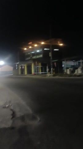 The video shows a dark, outdoor scene at night. A person is walking along a paved path, next to a road. There are streetlights visible, and the area appears to be urban or suburban, possibly near a commercial area with buildings and illuminated signs in the background. The camera is tilted, making it difficult to discern specific details, but the overall impression is of a nighttime journey through an illuminated area.