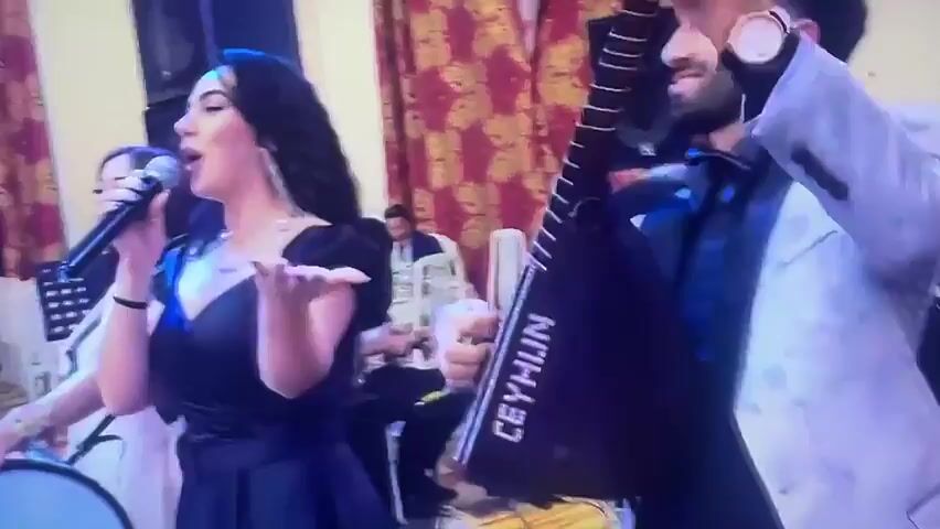 The video shows a live music performance at an indoor event. In the foreground, a woman wearing a black off-the-shoulder dress sings into a microphone. To her right, a man in a light-colored suit and black bowtie plays a stringed instrument that appears to be a saz, with the word "CEYHUN" visible on it. Behind them, other musicians are visible, including a person playing a drum or darbuka on the far left, and several men in red vests playing woodwind instruments and drums further back. The setting includes patterned curtains, chairs, speakers, and musical equipment, suggesting a performance space like a banquet hall or event venue. The atmosphere appears lively, with the musicians actively engaged in their performance.