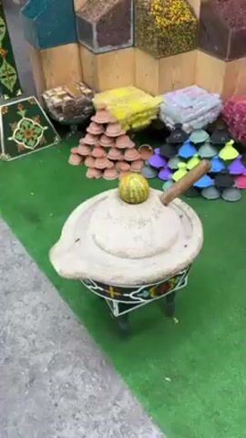 A close-up shot displays an outdoor market stand. The stand is decorated with a variety of objects, including stacks of small terracotta and plastic pyramids, as well as bags of seeds and other items. At the center of the frame is a traditional stone mill mounted on a decorated stand. The mill is light-colored and has a handle. A small melon rests on top of the mill. The ground around the stand is covered in artificial green turf. A person’s feet can be seen briefly in the background.