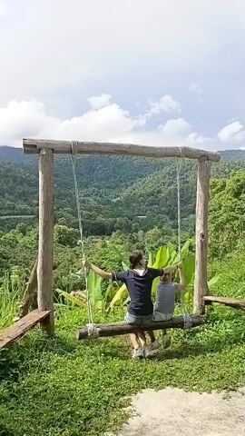 A wooden swing set with a thick log seat is set up on a grassy hillside overlooking a lush, green valley with rolling mountains in the background. Two people, one wearing a dark blue shirt and shorts, and the other a light gray shirt and shorts, are sitting on the swing, facing away from the camera, enjoying the view. They are gently swinging back and forth. The person in the dark blue shirt has their arms spread wide. The sky is partly cloudy. The overall scene depicts a serene and scenic outdoor setting.