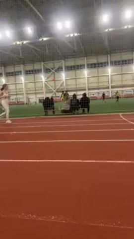 A woman in white athletic wear is running down a red indoor track. In the background, several people are congregated near the edge of a green indoor sports field. Some people can be seen playing on the field. The indoor space is lit by overhead lights. The camera is following the running woman from a side angle.
