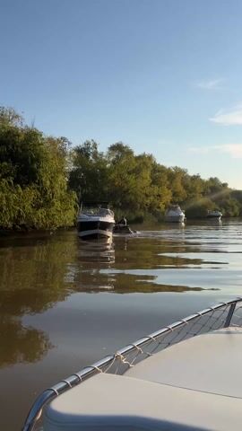 The video shows a boat moving along a narrow, reflective river. The river is surrounded by lush green trees on both banks. Three other boats are stationary in the water ahead. The sun shines brightly in the distance, almost directly in front of the boat's path, causing a glare in the video. The camera is positioned on the boat, looking forward, with the boat's white bow and metal railing visible.