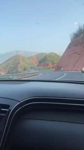 The video shows a view from inside a car looking out the windshield at a winding road through a mountainous landscape. The road curves to the right, with guardrails on the left side and a steep, reddish-brown mountain slope on the right. In the distance, rolling green hills with patches of red earth are visible, interspersed with trees and some buildings. The sky is clear and blue. A person on a motorcycle is seen driving on the road ahead, moving away from the camera. The dashboard of the car is visible in the foreground.