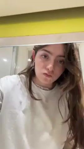 The video shows a young woman with long, wavy brown hair, wearing a white t-shirt and jewelry, in a bathroom setting. She's standing in front of a mirror with a yellow-colored top edge. In her hands, she holds a cotton pad, which she sprays with a liquid from a blue and white spray bottle. Following this, she uses the moistened cotton pad to clean around her ear, specifically near her earrings. The background includes a shower and a closed white door. The focus is primarily on the woman and her actions related to cleaning her ear.