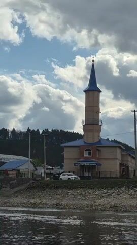 The video shows a building with a tall tower next to a river. The building has a blue roof and a light-colored brick exterior. A white car is parked in front of it. A sign above the entrance reads “Мечеть имени Мусы.” The tower is taller than the rest of the building and has a blue pointed roof.  The river is in the foreground, and there is a rocky bank.  The sky is cloudy with patches of blue.  Later in the video, power poles and tall pine trees can be seen along the riverbank.